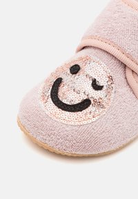 Zapatilla de bebé en suave color rosa con un parche de lentejuelas que muestra una carita sonriente guiñando un ojo en la parte frontal.
