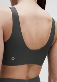 Tumši pelēks sporta krūšturis ar platām siksnām, izgriezumu aizmugurē un gludu auduma tekstūru. Mazs logotipa elements tuvumā jostasvietai.