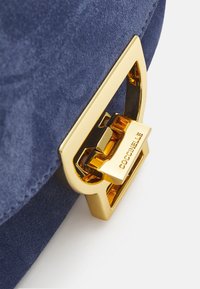 Marinblå mockaväska med spänne i guldfärgat metall med ingraverat varumärke, som visar en mjuk textur och ren geometrisk design.