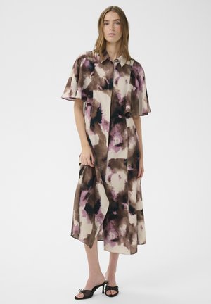 Nainen, joka pukeutunut löysään, polvenpituisen mekon, jossa on leveät hihat, ja joka on koristeltu ruskealla, mustalla ja violetilla abstraktilla kuviolla. Hänellä on myös mustat korkokengät.