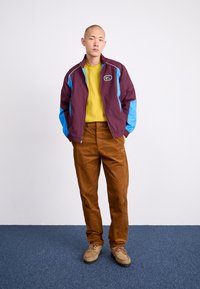 Geacă tip windbreaker cu panouri mov și albastre, cămașă galbenă, pantaloni din catifea groasă maro și pantofi bej pe un fundal de covor albastru.