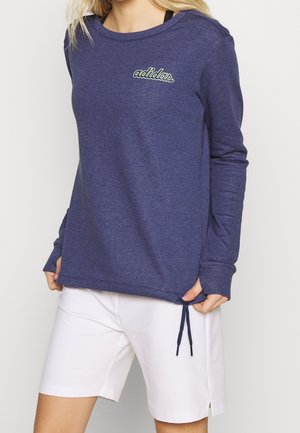 Kobieta nosząca granatową, długorękawową bluzę Adidas z białymi szortami, stojąca z rękami trzymającymi dół bluzy.