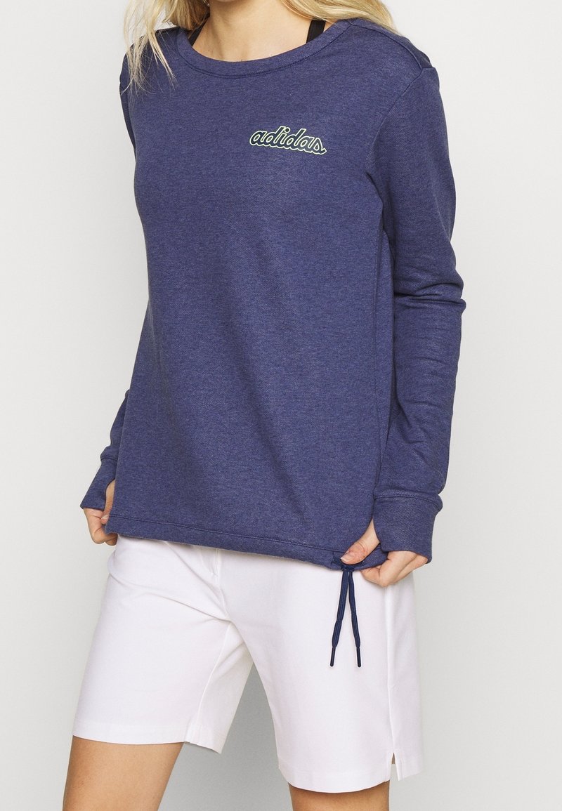 Femme portant un sweat-shirt à manches longues Adidas bleu marine avec un short blanc, debout, les mains tenant l'ourlet du sweat-shirt.