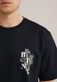 Fekete pamut póló, lila íriszekből álló virágmintázattal és fehér "BTNC" felirattal. Kerek nyakú, rövid ujjú kialakítás.
