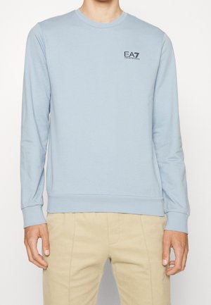Man draagt lichtblauwe longsleeve met EA7 Emporio Armani-logo op de borst en beige broek, staand voor een witte achtergrond.