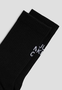 Schwarze gerippte Socken mit weißem Textbranding. Das Material ist glatt und betont eine enge Passform sowie ein klassisches Design. Zwei Paar sind gefaltet abgebildet.
