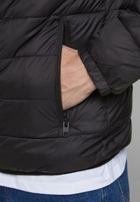 Zwarte pufferjack met een ritszak aan de zijkant, met een gladde, glanzende textuur en een quiltpatroon. Gelaagd over een witte shirt.