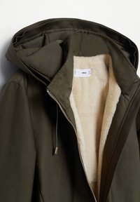 Veste à capuche vert foncé avec doublure en fausse peau de mouton beige et fermeture éclair argentée partiellement ouverte, dévoilant l'étiquette de la marque à l'intérieur.