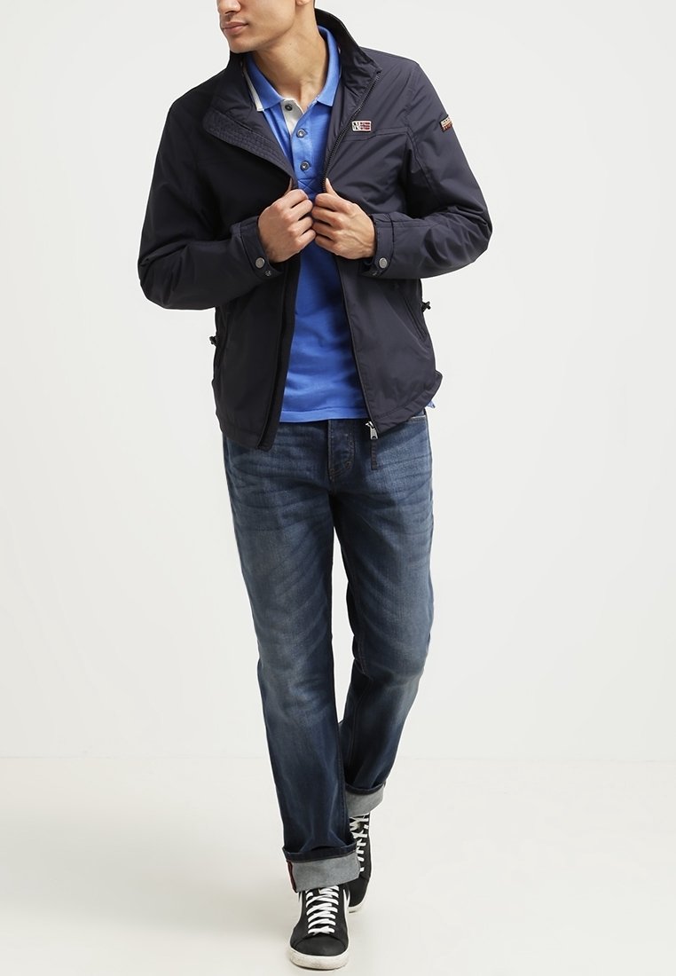 Marinblå lätt jacka med dragkedja, ståkrage och logodetalj, bärs över en blå polo och matchas med mörka jeans och sneakers.