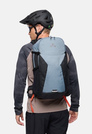 Persoon met een zwarte helm, zonnebril en een tweekleurige grijs-zwarte rugzak met oranje banden, gezien van achteren tegen een witte achtergrond.