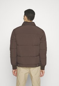 Brązowa kurtka puchowa z prążkowanym kołnierzem, z poziomym przeszyciem i elastycznymi mankietami. Noszona z oliwkowymi spodniami, widok od tyłu.