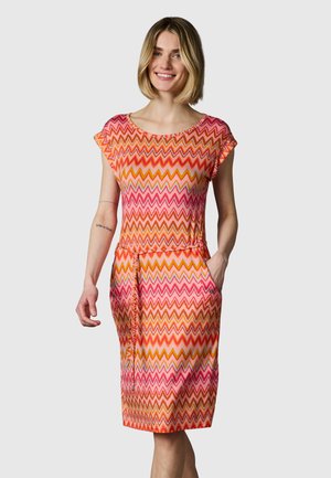 Donna sorridente che indossa un vestito al ginocchio con motivo a zigzag rosa, arancione e rosso, maniche corte e tasche laterali, in piedi davanti a uno sfondo semplice.