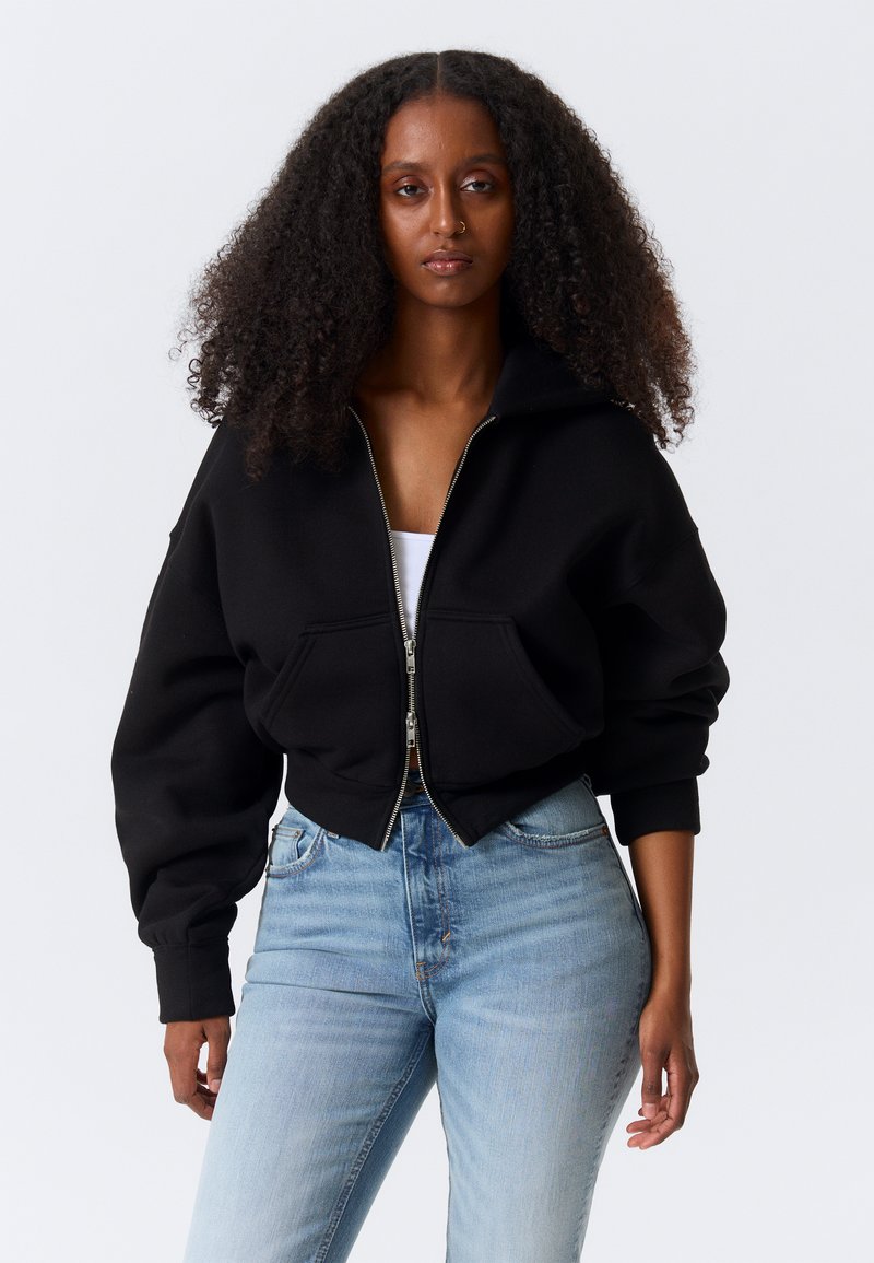 Γυναίκα με μακριά, μπούκλες μαλλιά, που φοράει μαύρο μπουφάν με φερμουάρ πάνω από λευκό τοπ και ανοιχτόχρωμα μπλε τζιν, στέκεται μπροστά σε απλό φόντο.