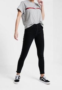 Camiseta gris de manga corta con una franja color burdeos, combinada con jeans ajustados negros que tienen franjas laterales. Zapatillas negras completan el conjunto.