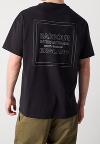 Černé bavlněné tričko s bílým grafickým textovým designem na zadní straně, který uvádí "BARBOUR INTERNATIONAL SOUTH SHIELDS ENGLAND." Krátké rukávy.