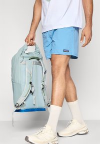 Ανοιχτό μπλε σακίδιο με γκρι ιμάντες και πράσινη λεπτομέρεια, κατασκευασμένο από ανθεκτικό ύφασμα. Φοριέται με αντιστοιχούντες μπλε σορτς και μπεζ παπούτσια.