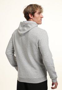 Giovane uomo con capelli biondi che indossa una felpa con cappuccio grigia chiara, girato leggermente per mostrare il retro e il profilo laterale.