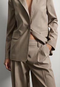 Costume beige sur mesure avec blazer à bouton unique et revers crantés, associé à un pantalon à jambes larges. Tissu lisse avec une texture subtile.