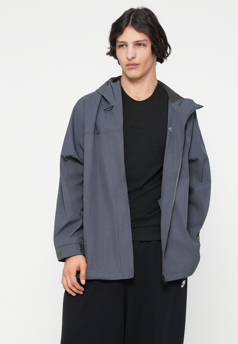 Grijze waterdichte jacket met een capuchon, ritssluiting, handzakken en verstelbare manchetten. Gecombineerd met een zwart T-shirt en losse zwarte broek.