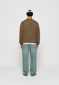 Brun strikket genser med avslappet passform, ribbestrikket nederkant og senkede skuldre, kombinert med lyseblå denimjeans og en beige caps.