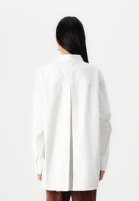 White long-sleeve shirt with a back vent, buttoned cuffs, and a straight-cut silhouette, made from smooth fabric.