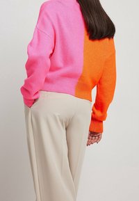 Pull color-blocké avec du rose à gauche et de l'orange à droite, fabriqué en matière tricotée douce. Porté avec un pantalon beige. Manches longues, coupe décontractée.