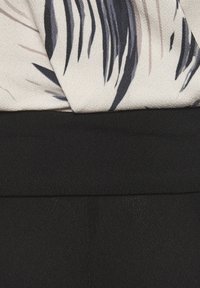 Licht stof met een bloemmotief, voorzien van beige en zwarte bladeren, gecombineerd met een effen zwarte textured tailleband, wat een gelaagd uiterlijk creëert.