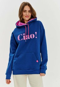 Modrý svetr s růžovou kapucí a šňůrkami, na přední straně s nápisem "Ciao!" v tučných růžových písmenech a s přední kapsou.