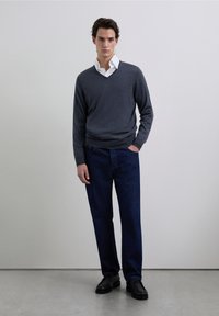 Szary sweter z dekoltem w serek na białą koszulę z kołnierzykiem, zestawiony z ciemnoniebieskimi dżinsami i czarnymi butami. Prosty, dopasowany krój o gładkiej fakturze.