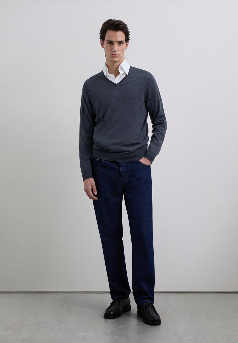 Szary sweter z dekoltem w serek na białą koszulę z kołnierzykiem, zestawiony z ciemnoniebieskimi dżinsami i czarnymi butami. Prosty, dopasowany krój o gładkiej fakturze.