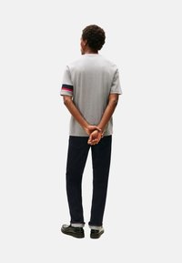 Camiseta gris de manga corta con acentos a rayas rojas y marino en las mangas, combinada con jeans de denim azul oscuro y zapatos negros.