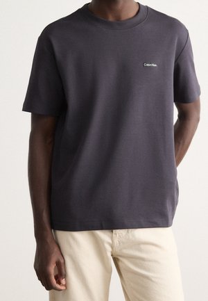 Homme portant un T-shirt Calvin Klein gris foncé à manches courtes et un pantalon beige clair, mains détendues le long du corps.