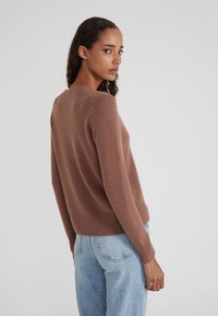 Frau mit lockigem Haar, die einen braunen Langarmpullover und hellblaue Jeans trägt, leicht gedreht und über ihre Schulter blickend.