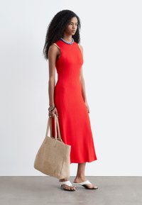 Femme aux cheveux bouclés portant une robe midi rouge côtelée sans manches, un collier bleu, des sandales blanches à talons, tenant un grand sac fourre-tout tissé beige.