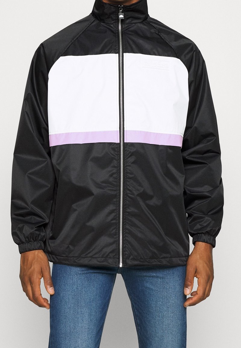 Schwarze und weiße Windjacke mit einem lila horizontalen Streifen, Reißverschluss, elastischen Bündchen und hohem Kragen. Hat eine glatte Textur.