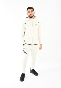 Chándal deportivo blanco con acentos negros, que incluye una sudadera con cremallera y pantalones ajustados. El modelo lleva zapatillas de correr negras.