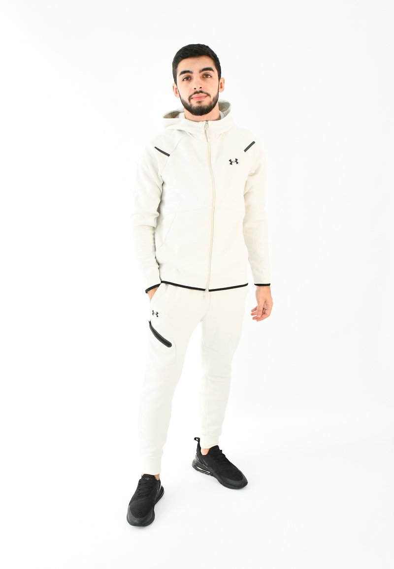 Chándal deportivo blanco con acentos negros, que incluye una sudadera con cremallera y pantalones ajustados. El modelo lleva zapatillas de correr negras.