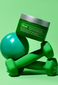 Grön Murad Retinal Resculpt Body Treatment jar med silverlock, placerad ovanför en grön träningsboll och två gröna hantlar, mot en grön bakgrund.