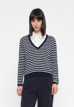 Young woman with straight dark hair wearing a white collared shirt under a navy and white striped V-neck sweater and dark trousers.
