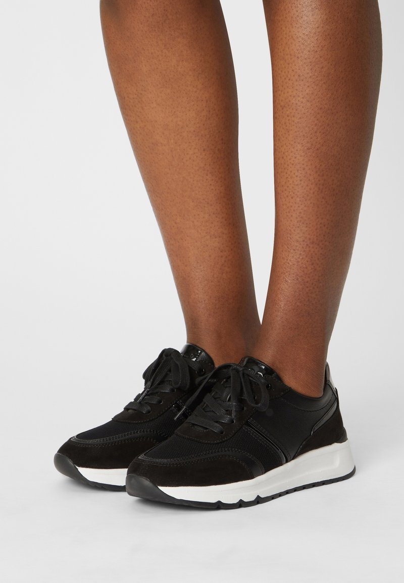 Jambes portant des baskets noires à lacets avec des semelles blanches, debout sur un sol gris clair uni avec un fond neutre.