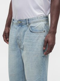 Personne portant un jean en denim bleu clair et un t-shirt blanc uni, debout les bras détendus le long du corps sur un fond uni.