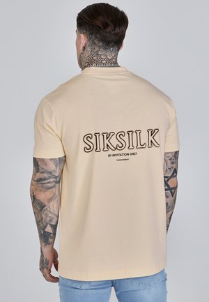 T-shirt beige à manches courtes avec col rond, présentant "SIKSILK BY INVITATION ONLY" en lettres majuscules au dos. Matière en coton doux.