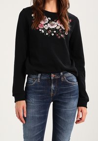 Sweatshirt noir avec broderie florale en rose, blanc et bordeaux. Associé à un jean en denim à lavage moyen. Texture douce, coupe décontractée.