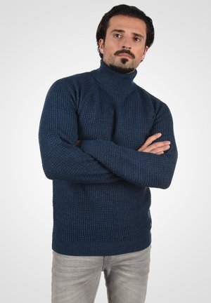Marineblauwe gebreide coltrui met ribbelstructuur, iets losse pasvorm en effen kleur ontwerp. Draagt met lichtgrijze broeken.