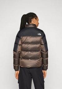 Bruine en zwarte pufferjack met een hoge kraag, voorzien van een doorgestikt ontwerp en logo op de achterkant. Gladde, synthetische stof, elastische manchetten.