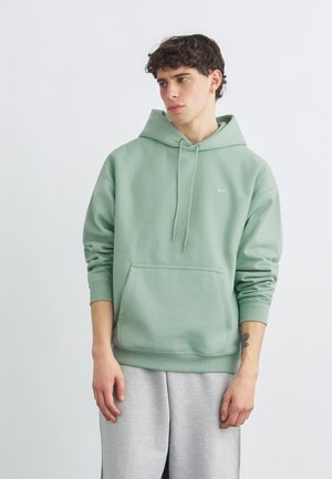 Νεαρός άνδρας με κοντά σγουρά μαλλιά, ντυμένος με ένα φούτερ πράσινου χρώματος σοδειάς και ανοιχτό γκρι φόρμα, στέκεται μπροστά σε ένα απλό ανοιχτό φόντο.