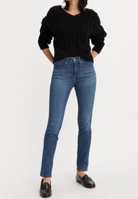 Suéter negro de punto trenzado, jeans ajustados azules y mocasines negros. Los jeans tienen una cintura alta y un desvanecimiento sutil. Estilo simple y clásico.