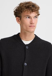 Cardigan nero a coste con scollatura tonda, due bottoni scuri e tessuto testurizzato. Camicia bianca visibile sotto.
