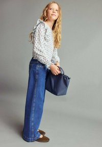 Blusa azzurro chiaro con maniche a volant, abbinata a jeans in denim a gamba larga e scarpe mocassino marroni; tiene in mano una piccola borsa blu navy.