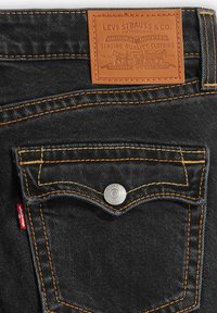 Jeans de mezclilla negra con costuras naranjas, una etiqueta de cuero con los detalles de la marca y un botón plateado en el bolsillo delantero.